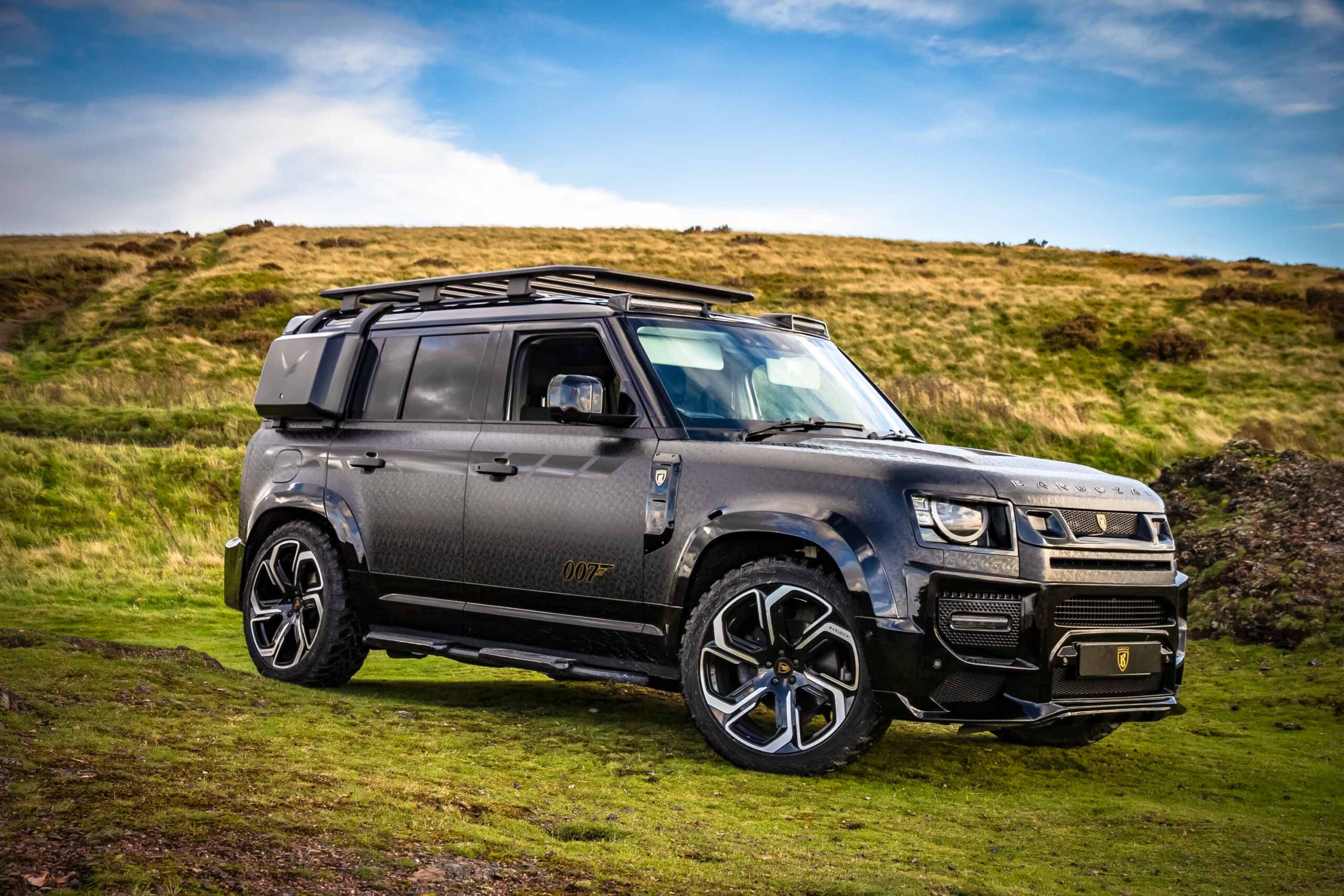 Front quarter view of a custom wrapped Land Rover Defender 110 L663 2020+. Shot outside near mountains. Fitted with Barugzai Sabre 22” alloy wheels, in Gloss Black Polished Face.