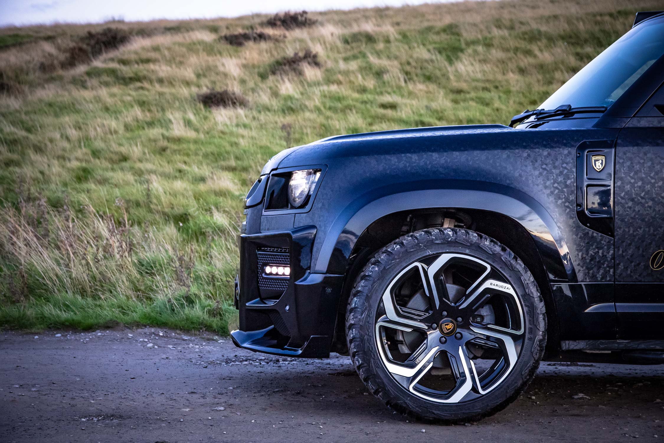 Side detail view of a custom wrapped Land Rover Defender 110 L663 2020+. Shot outside near mountains. Fitted with Barugzai Sabre 22” alloy wheels, in Gloss Black Polished Face.