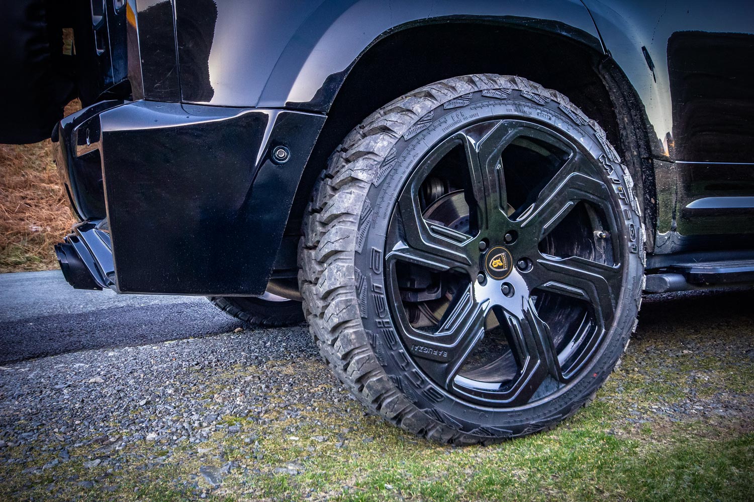 Rear quarter detail view of a custom wrapped Land Rover Defender 110 L663 2020+. Shot outside near mountains. Fitted with Barugzai Sabre 22” alloy wheels, in Gloss Black.