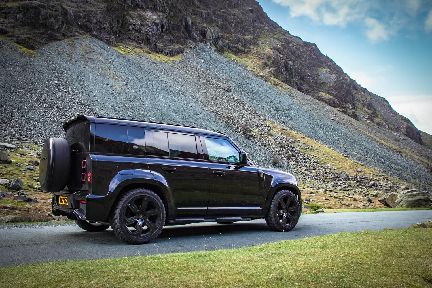 Rear quarter view of a custom wrapped Land Rover Defender 110 L663 2020+. Shot outside near mountains. Fitted with Barugzai Sabre 22” alloy wheels, in Gloss Black.