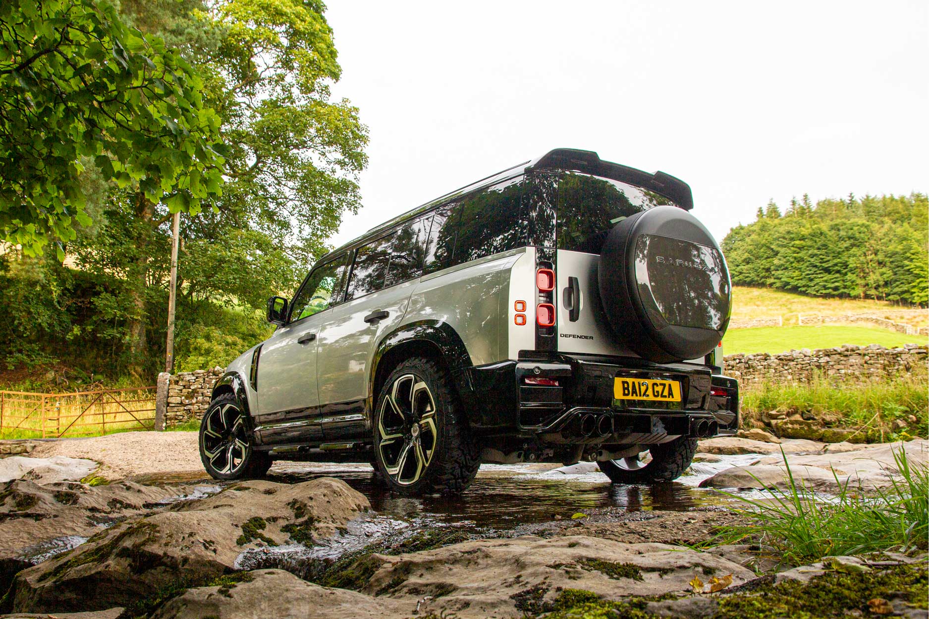Rear quarter view of a Silver Land Rover Defender 110 L663 2020+. Shot outside a hiking trail. Fitted with Barugzai Sabre 22” alloy wheels, in Gloss Black Polished Face.