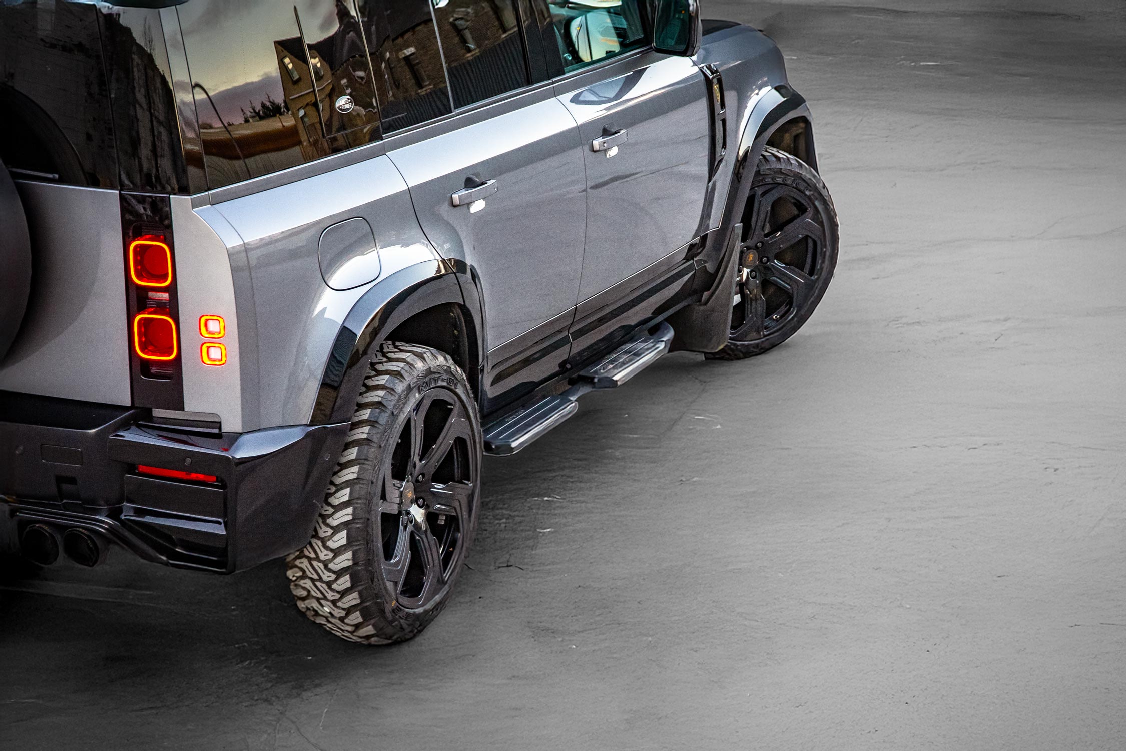Rear quarter detail view of a silver Land Rover Defender 110 L663 2020+. Shot outside an empty warehouse. Fitted with Barugzai Sabre 22” alloy wheels, in Gloss Black.