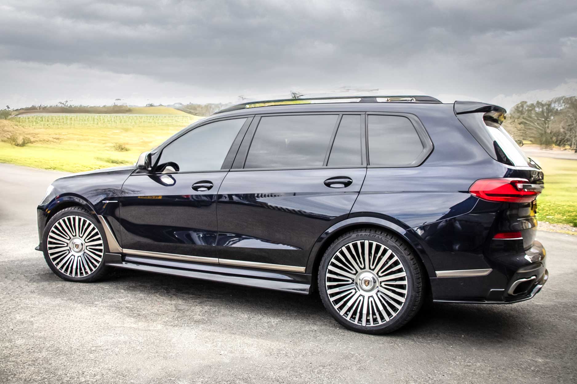 Rear quarter view of a Blue BMW X7 G07 2019+. Shot outside a rustic backdrop. Fitted with Barugzai Casanova 22/23” alloy wheels, in Gloss Black Polished Face.