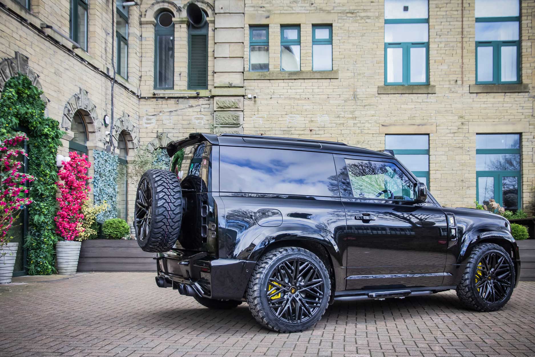 Rear quarter view of a Black Land Rover Defender 90 L663. Parked outside a restaurant Fitted with Barugzai Shuracan 22” alloy wheels, in Gloss Black.