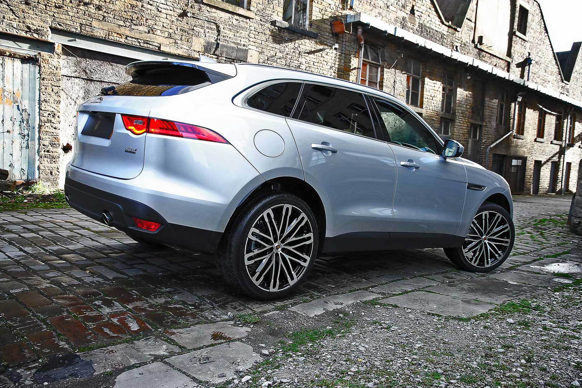 Rear quarter view of a Silver Jaguar F-Pace 2016+. Shot outside an old mill. Fitted with Barugzai Dawn 22” alloy wheels, in Gloss Black Polished Face.