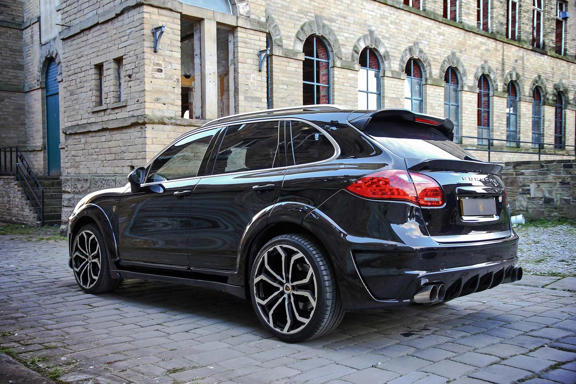 Rear quarter view of a Blue Porsche Cayenne 92A 2011-2018. Shot outside an old mill. Fitted with Barugzai Opium 22” alloy wheels, in Gloss Black Polished Face.