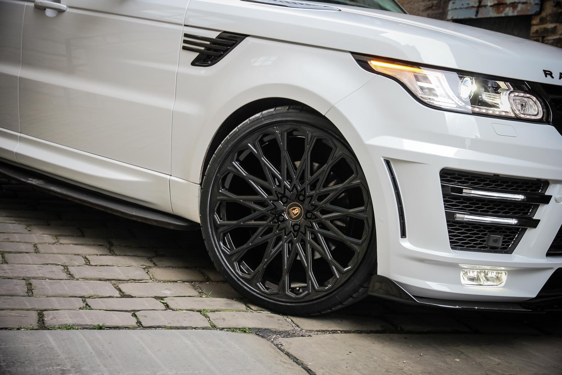 : Front quarter detail view of a White Range Rover Sport L494 2013-2017. Shot outside an old mill. Fitted with Barugzai Majestic 24” alloy wheels, in Gloss Black.