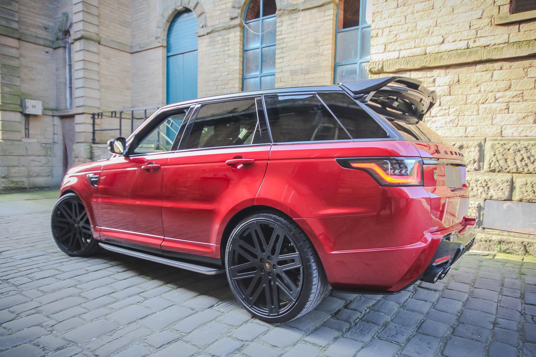 Rear quarter view of a Red Range Rover Sport L494 2018-2022. Shot outside an old mill. Fitted with Barugzai EXEL 24” alloy wheels, in Gloss Black.