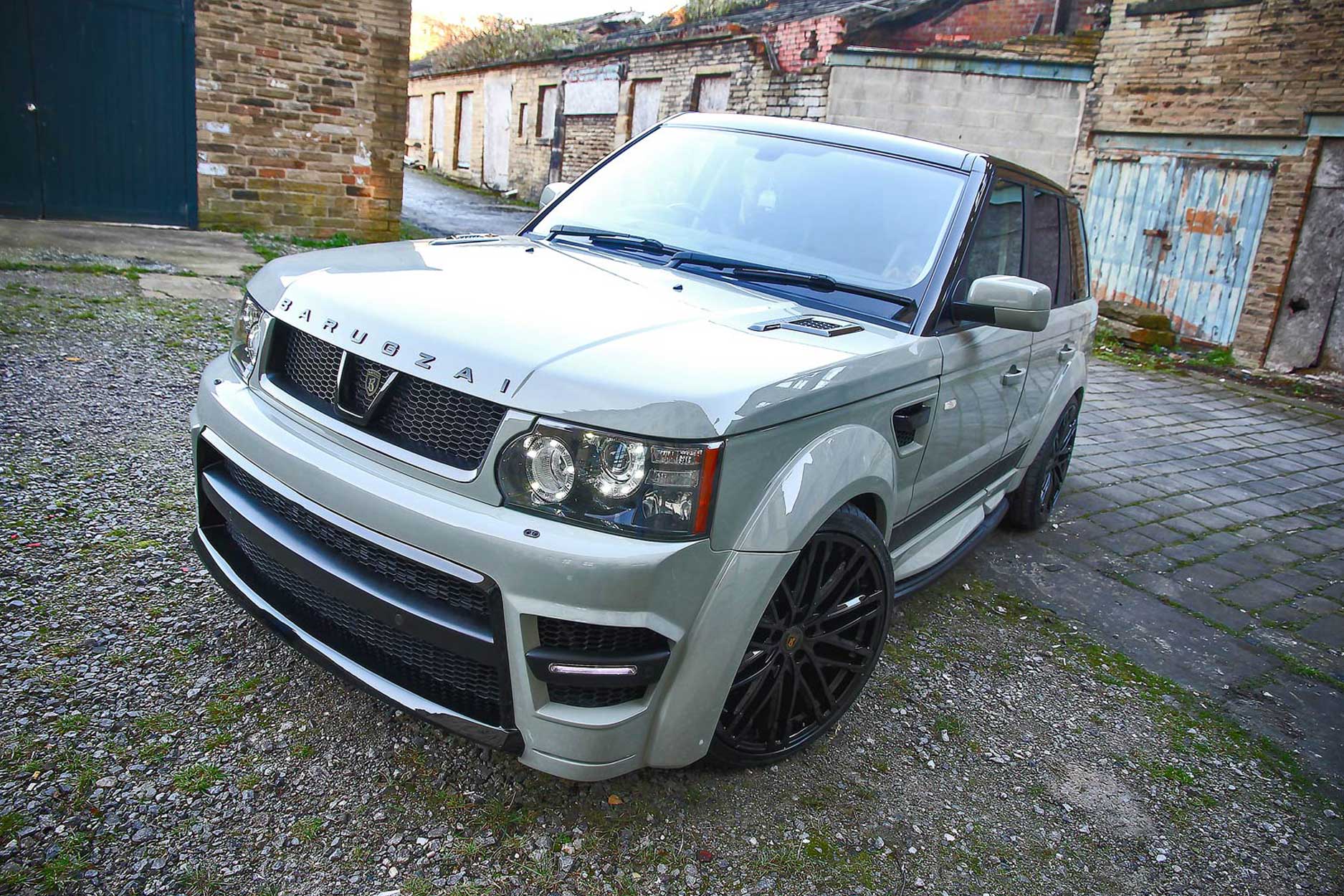 Front quarter view of a Nardo Grey Range Rover Sport L320 2009-2012. Shot outside an old mill. Fitted with Barugzai Dawn 22” alloy wheels, in Gloss Black.