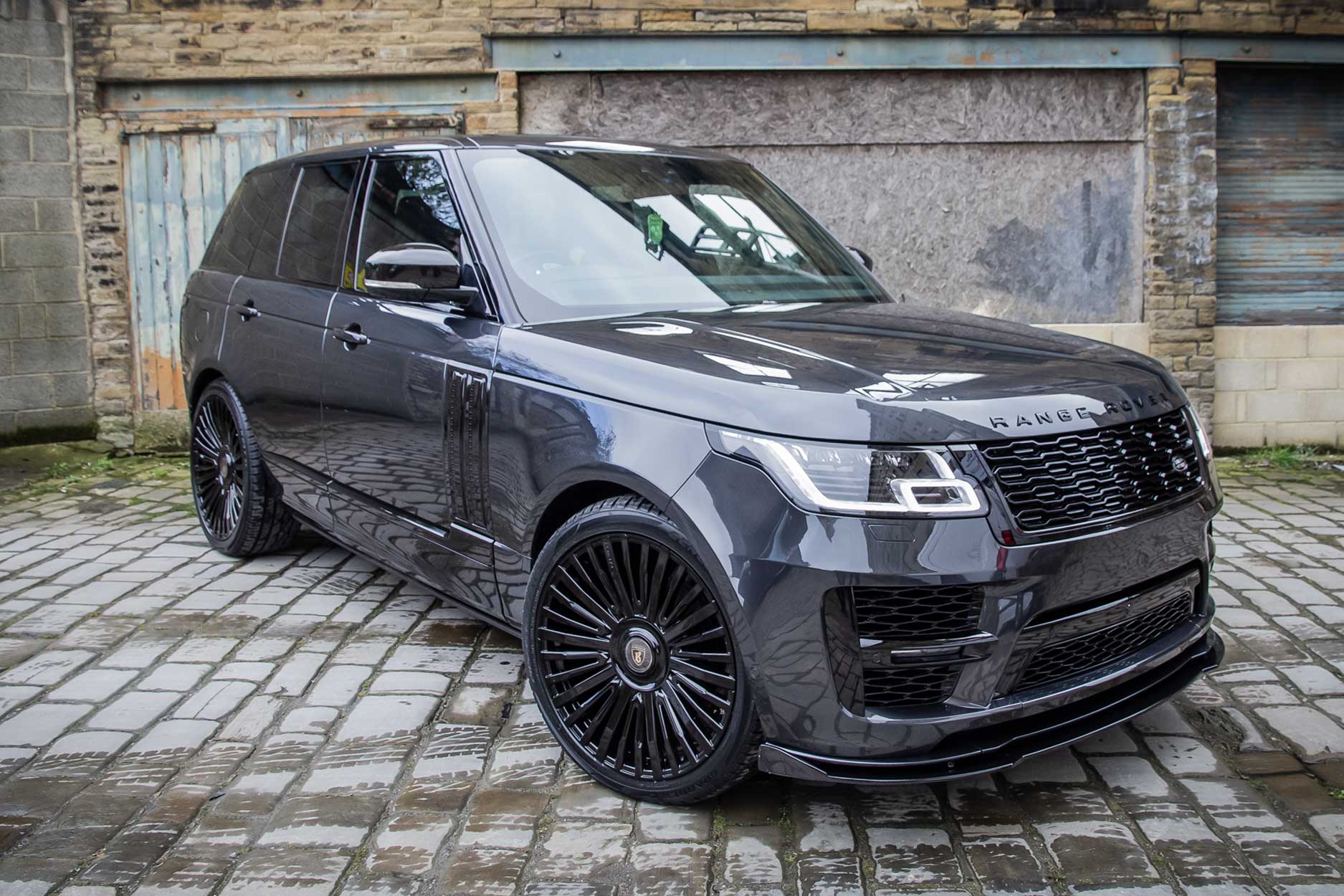 Front quarter view of a Carpathian Grey Range Rover Vogue L405 SVO style 2018-2021. Shot outside an old mill. Fitted with Barugzai Casanova 22/23” alloy wheels, in Gloss Black