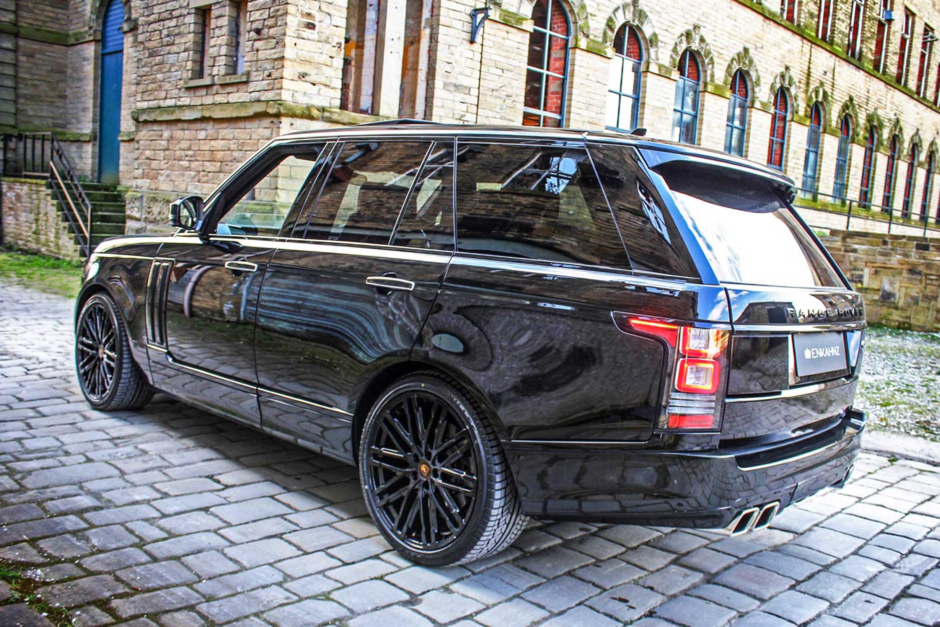 : Rear quarter view of a Black Range Rover L405 2013-2017. Shot outside an old mill. Fitted with Barugzai Dawn 22” alloy wheels, in Gloss Black