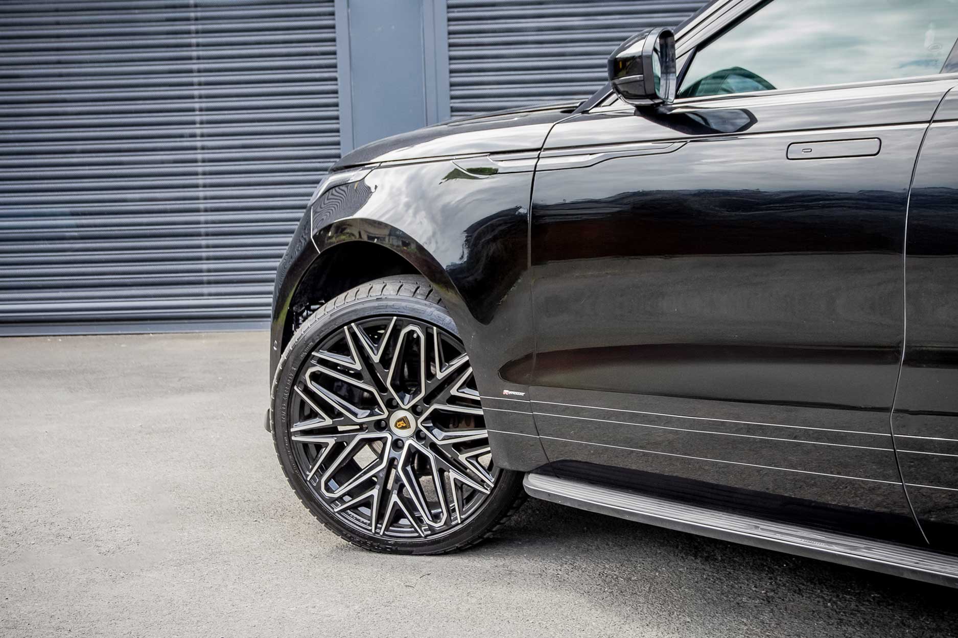 Side quarter detail view of a Black Range Rover Velar 2017+. Parked outside a building with a shutter. Fitted with Barugzai Shuracan 22” alloy wheels, in Gloss Black Polished Face.