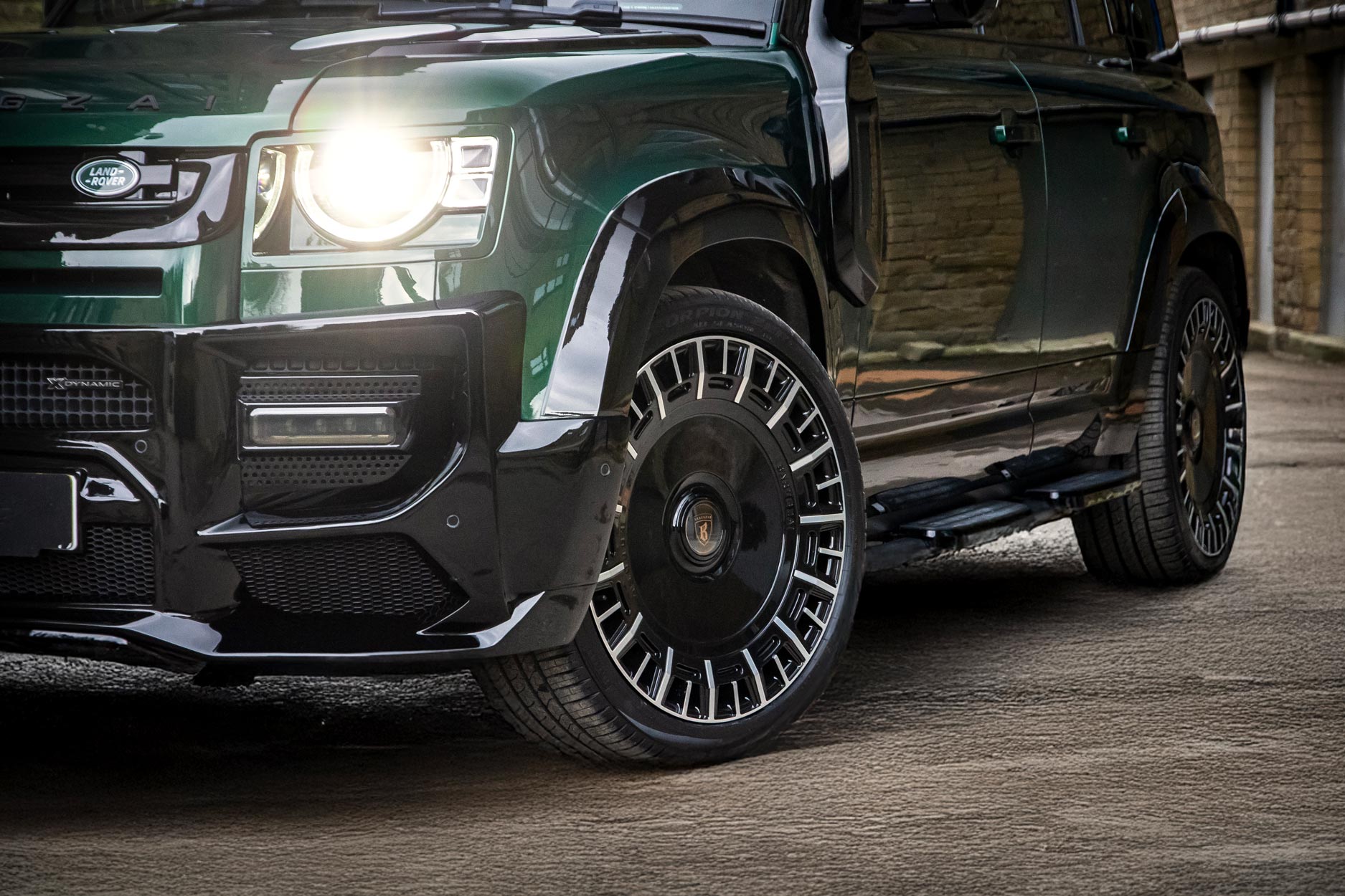 Front quarter detail view of a Green Land Rover Defender 110 L663 2020+. Shot outside an old mill. Fitted with Barugzai Monoblok 23” alloy wheels, in Gloss Black Polished Face.