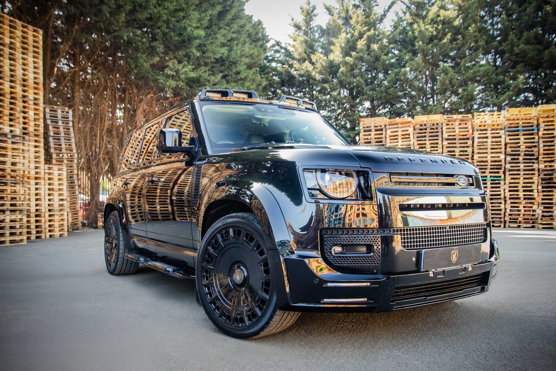 Front quarter view of a Black Land Rover Defender 110 L663 2020+. Shot outside pallet warehouse. Fitted with Barugzai Monoblok 23” alloy wheels, in Gloss Black.