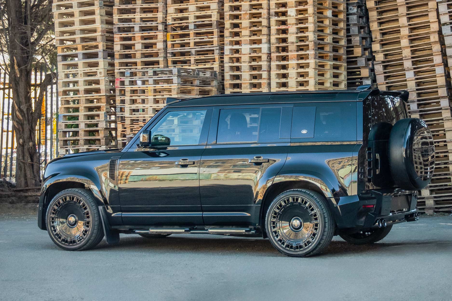 Side quarter view of a Black Land Rover Defender 110 L663 2020+. Shot outside pallet warehouse. Fitted with Barugzai Monoblok 23” alloy wheels, in Gloss Black.