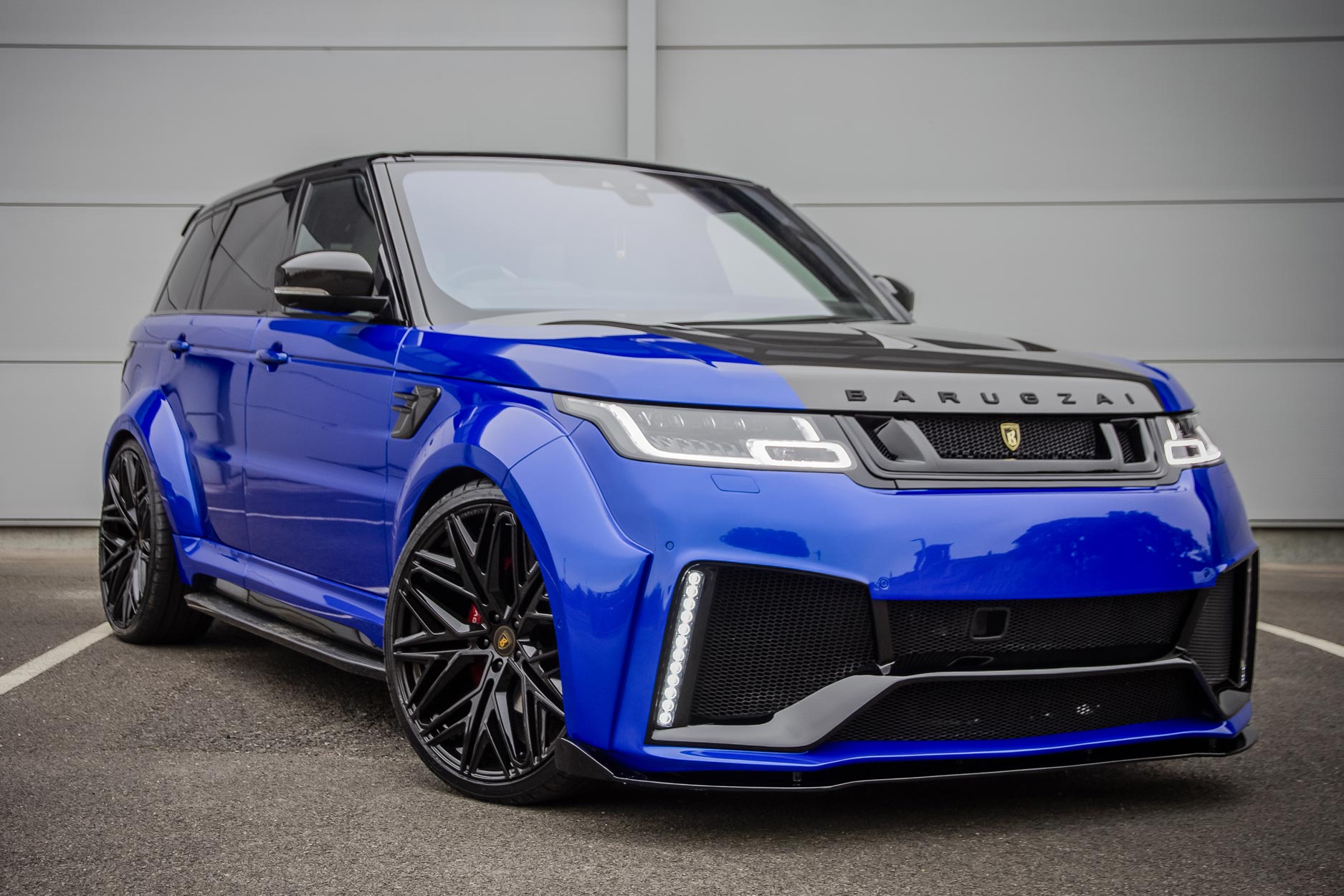Front quarter view of a Blue Range Rover Sport L494 2018-2022. Parked on a road with a grey background. Fitted with Barugzai Shuracan 24” alloy wheels, in Gloss Black.