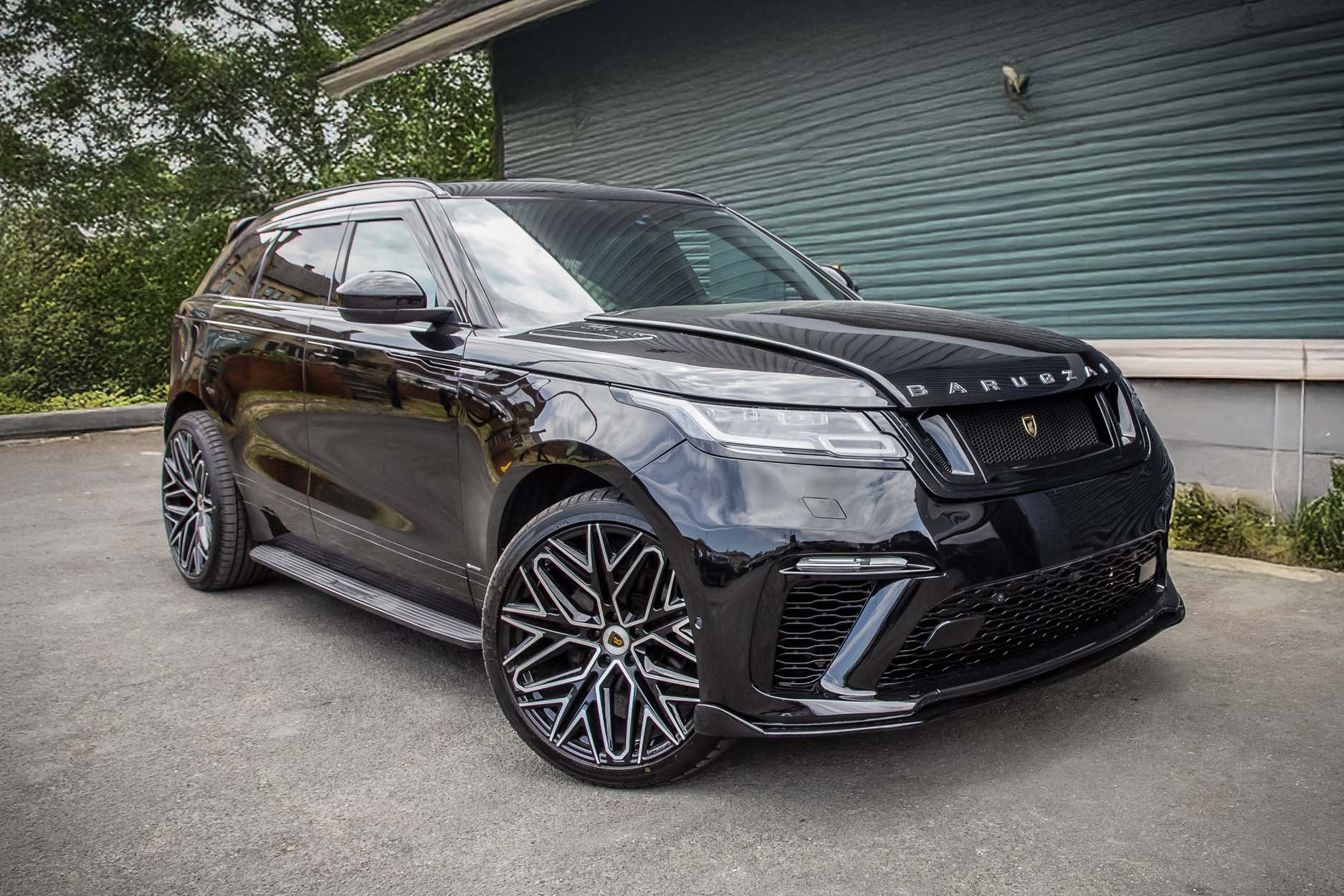 Front quarter view of a Black Range Rover Velar 2017+. Parked outside a building with a shutter. Fitted with Barugzai Shuracan 22” alloy wheels, in Gloss Black Polished Face.