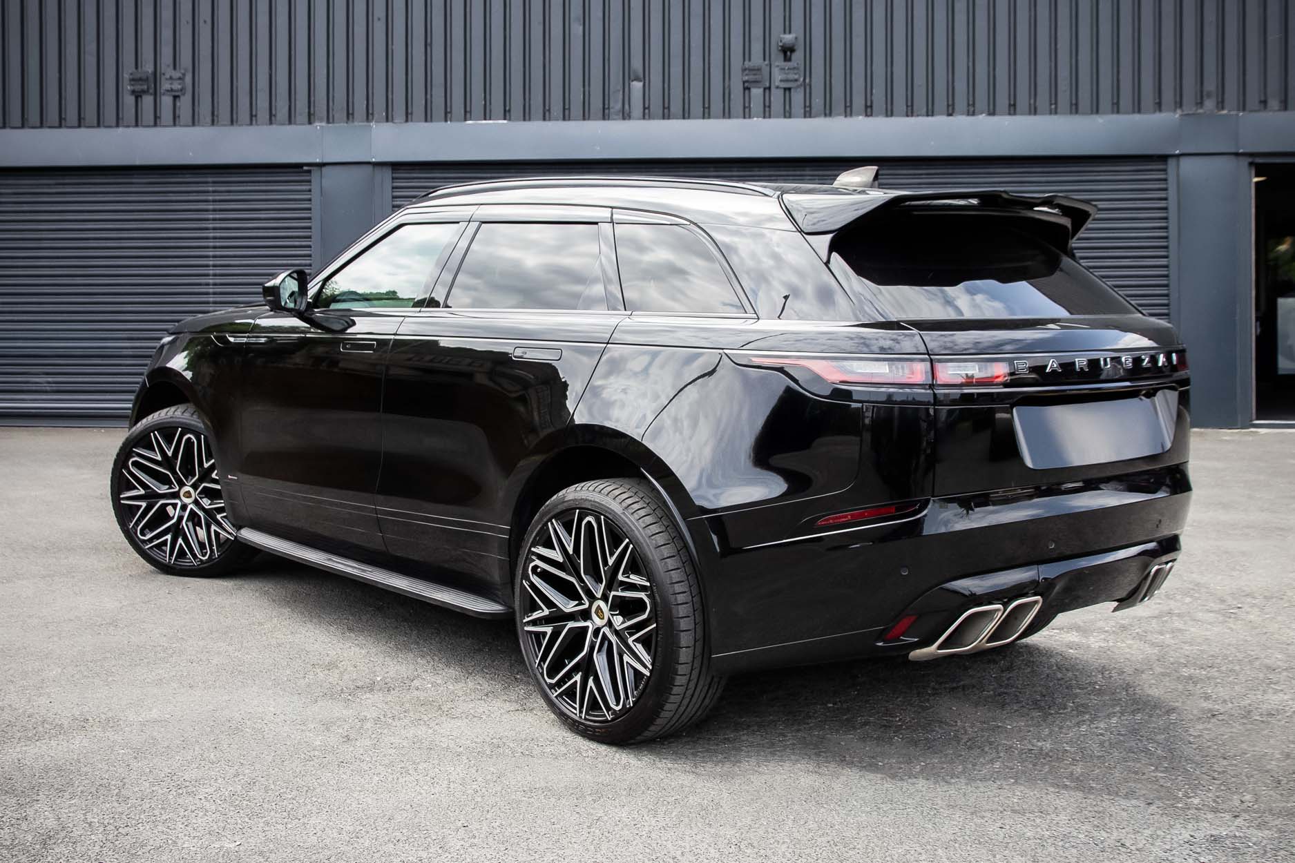 Rear quarter view of a Black Range Rover Velar 2017+. Parked outside a building with a shutter. Fitted with Barugzai Shuracan 22” alloy wheels, in Gloss Black Polished Face.