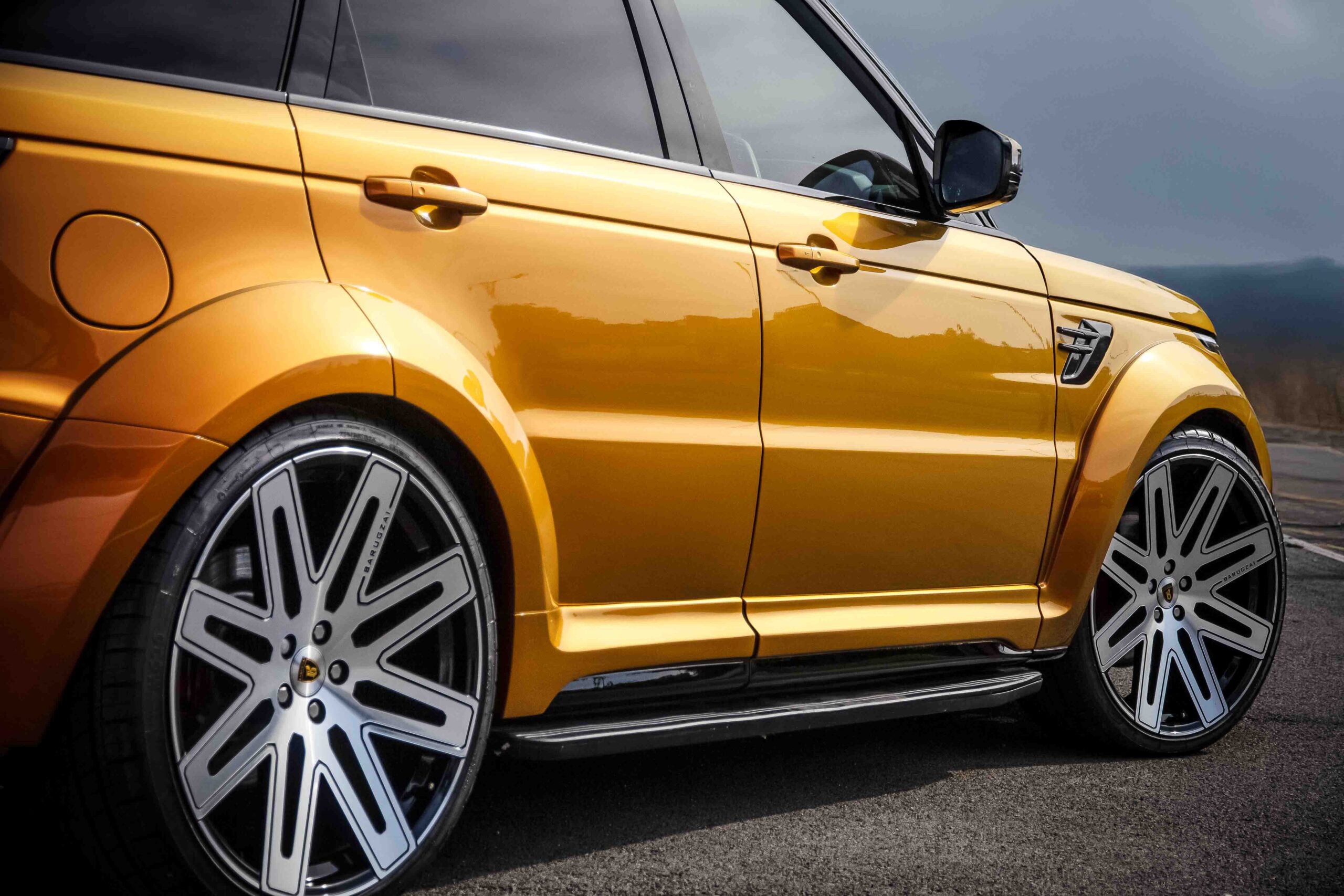 Side quarter detail view of an Orange Range Rover Sport L494 2018-2022. Shot outside a scenic backdrop. Fitted with Barugzai EXEL 24” alloy wheels, in Gloss Black Polished Face.