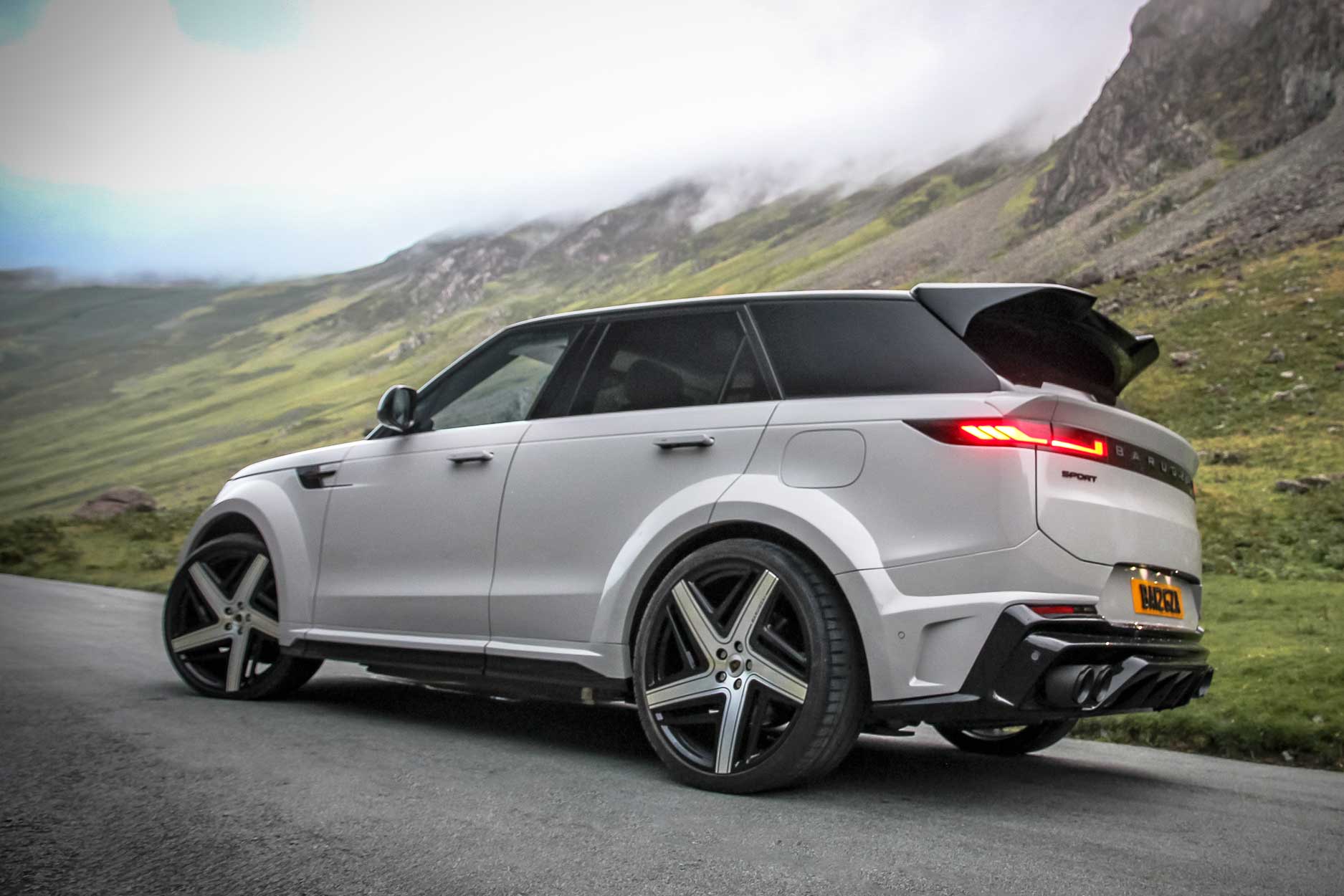 Rear quarter view of a Nardo Grey Range Rover Sport 2023+. Parked outside a mountain scenery. Fitted with Barugzai Eleven 24” alloy wheels, in Gloss Black Polished Face.