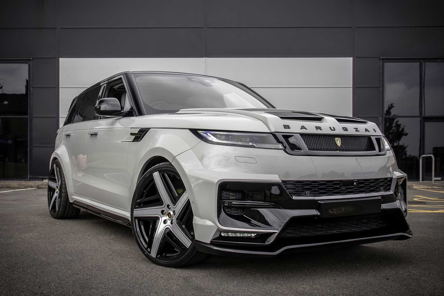 Front quarter view of a Nardo Grey Range Rover Sport 2023+. Parked on a road with a grey background. Fitted with Barugzai Eleven 24” alloy wheels, in Gloss Black Polished Face.
