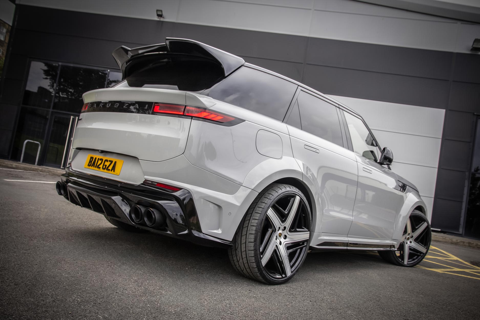 Rear quarter view of a Nardo Grey Range Rover Sport 2023+. Parked on a road with a grey background. Fitted with Barugzai Eleven 24” alloy wheels, in Gloss Black Polished Face.