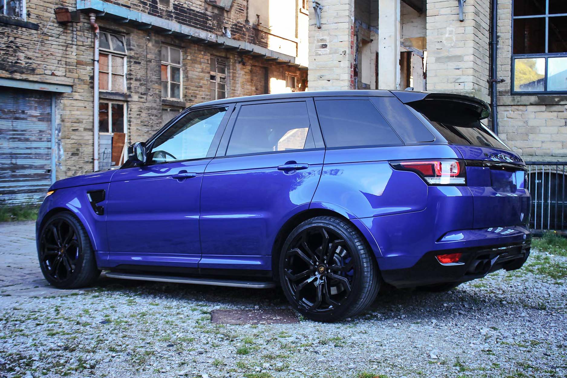 Rear quarter view of a Blue Range Rover Sport SVR L494 2013-2017. Shot outside an old mill. Fitted with Barugzai Opium 22” alloy wheels, in Gloss Black